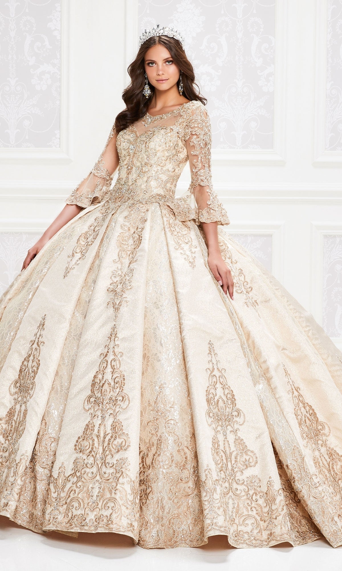 A young woman stands indoors wearing the Princesa PR12004 Quinceañera Dress, featuring gold embroidery, a full skirt, elegant bell sleeves, and a jeweled tiara.